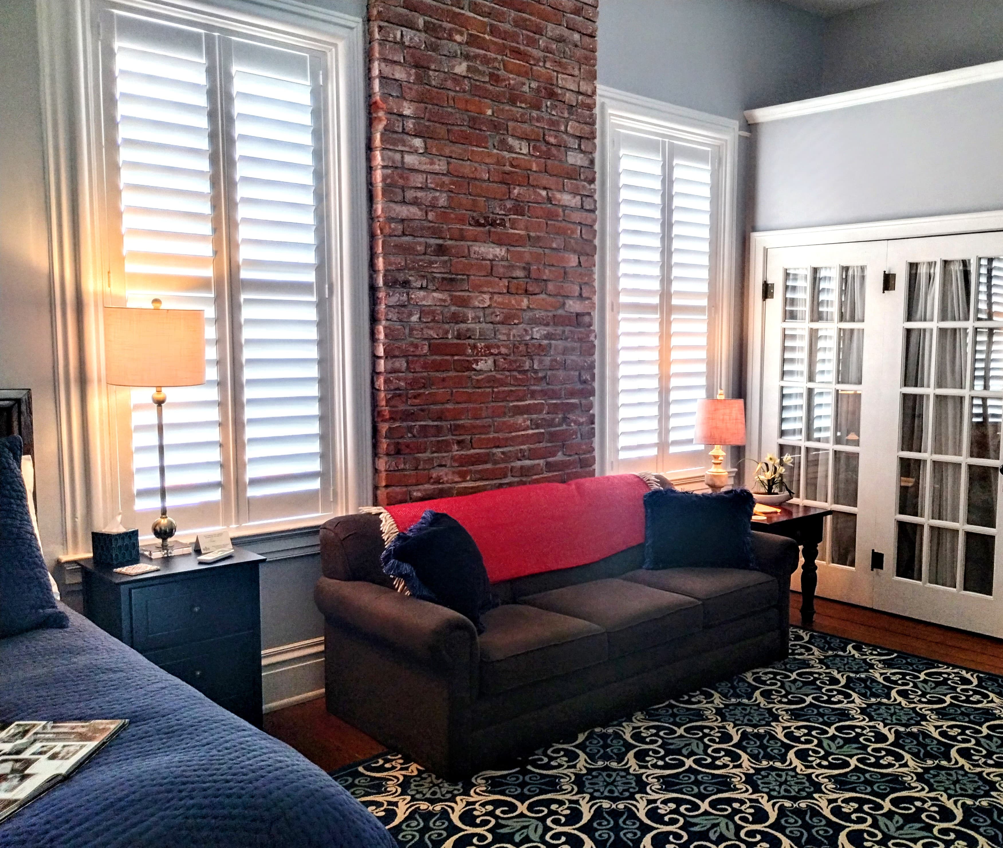 A sitting area with a dark couch, white shuttered windows, a brick accent wall, and a patterned rug.