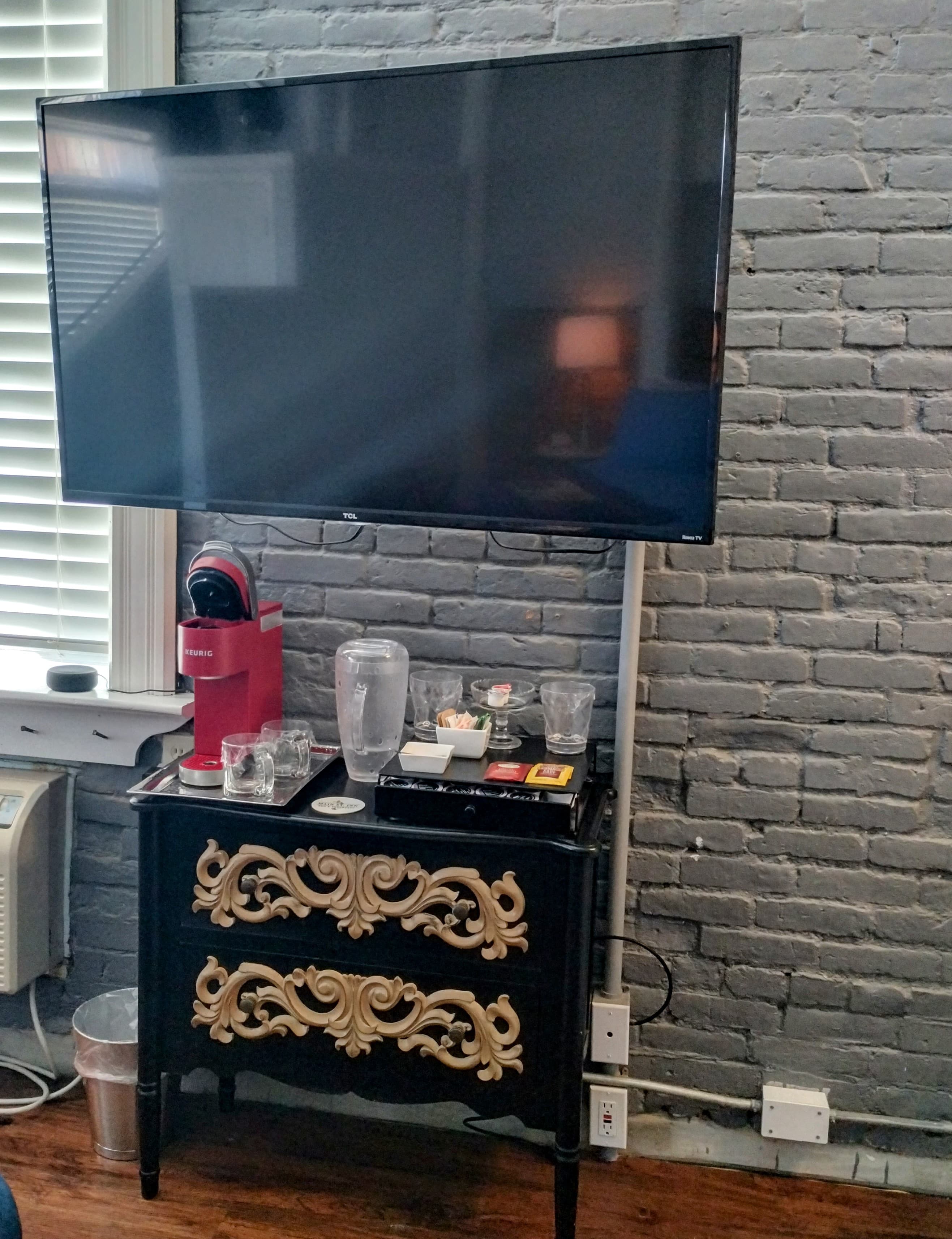 A flat-screen TV mounted above a black dresser with gold accents, against a gray brick wall.