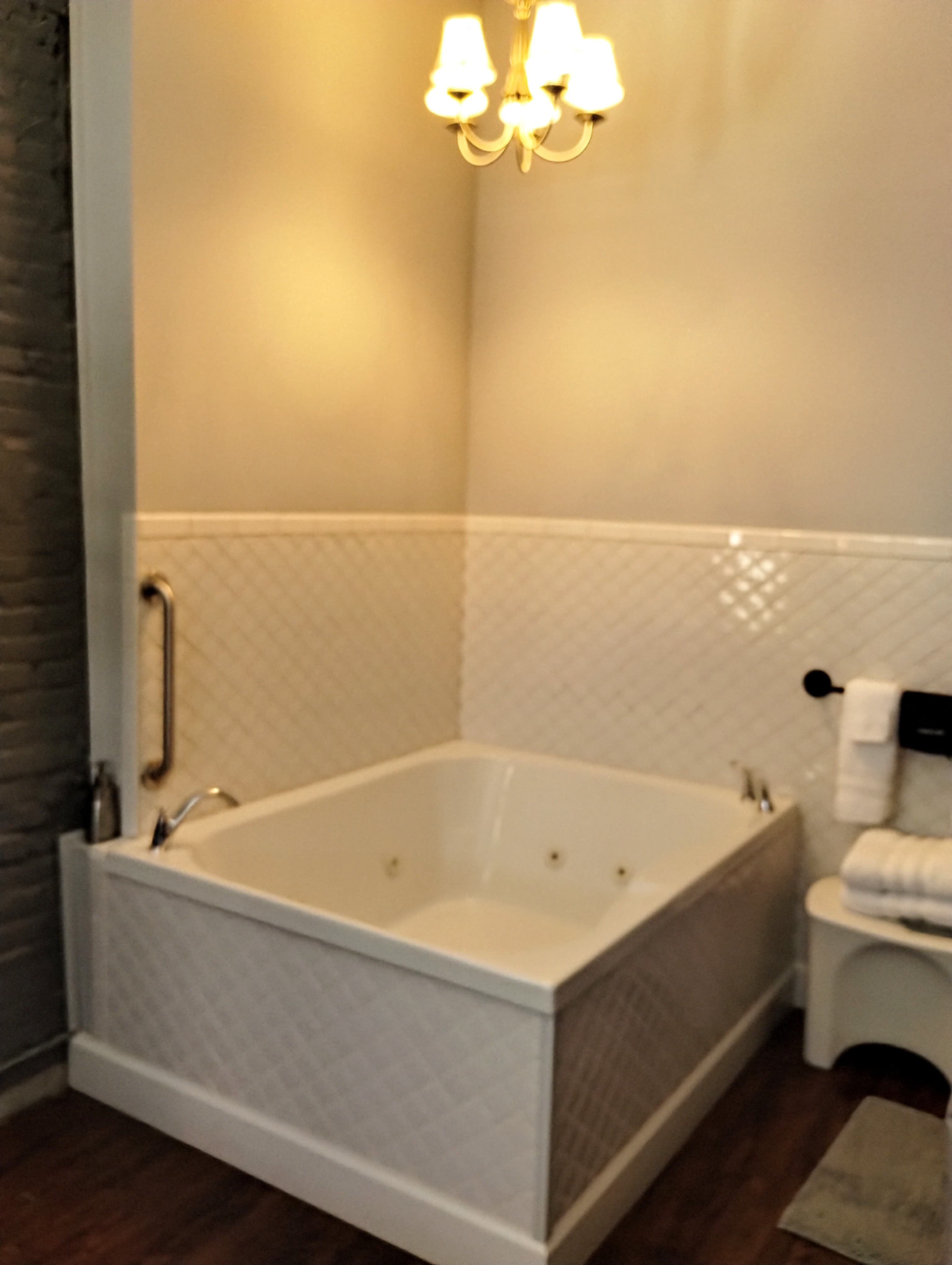 A bathroom with a white square jacuzzi tub, white tiled walls, and a chandelier.