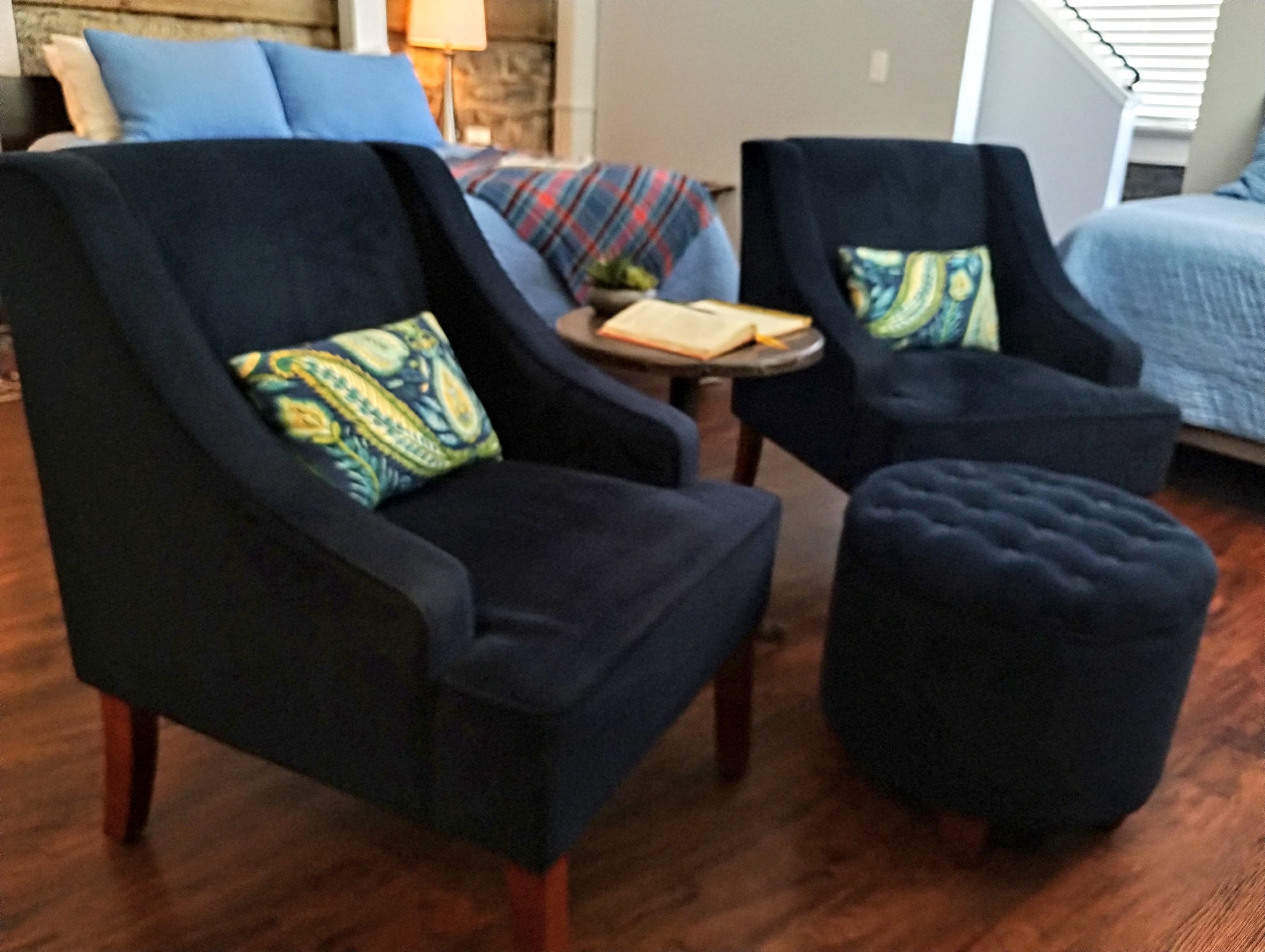 A cozy seating area with two dark blue armchairs, each with a patterned green and yellow pillow, a small round wooden table with a book, and a tufted black ottoman on a polished wood floor.