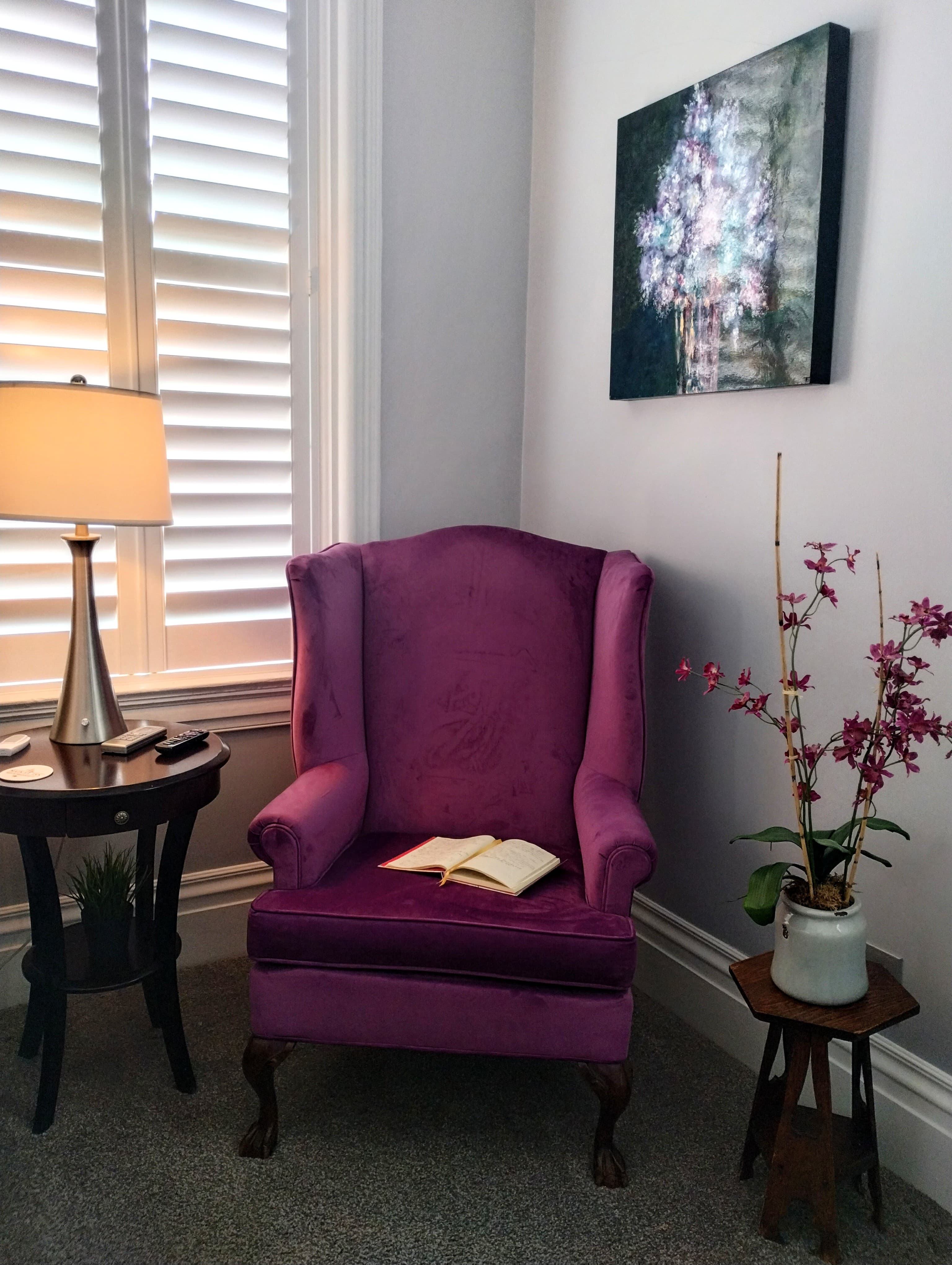 A cozy corner with a purple wingback armchair with a book on the seat, next to a window with blinds.