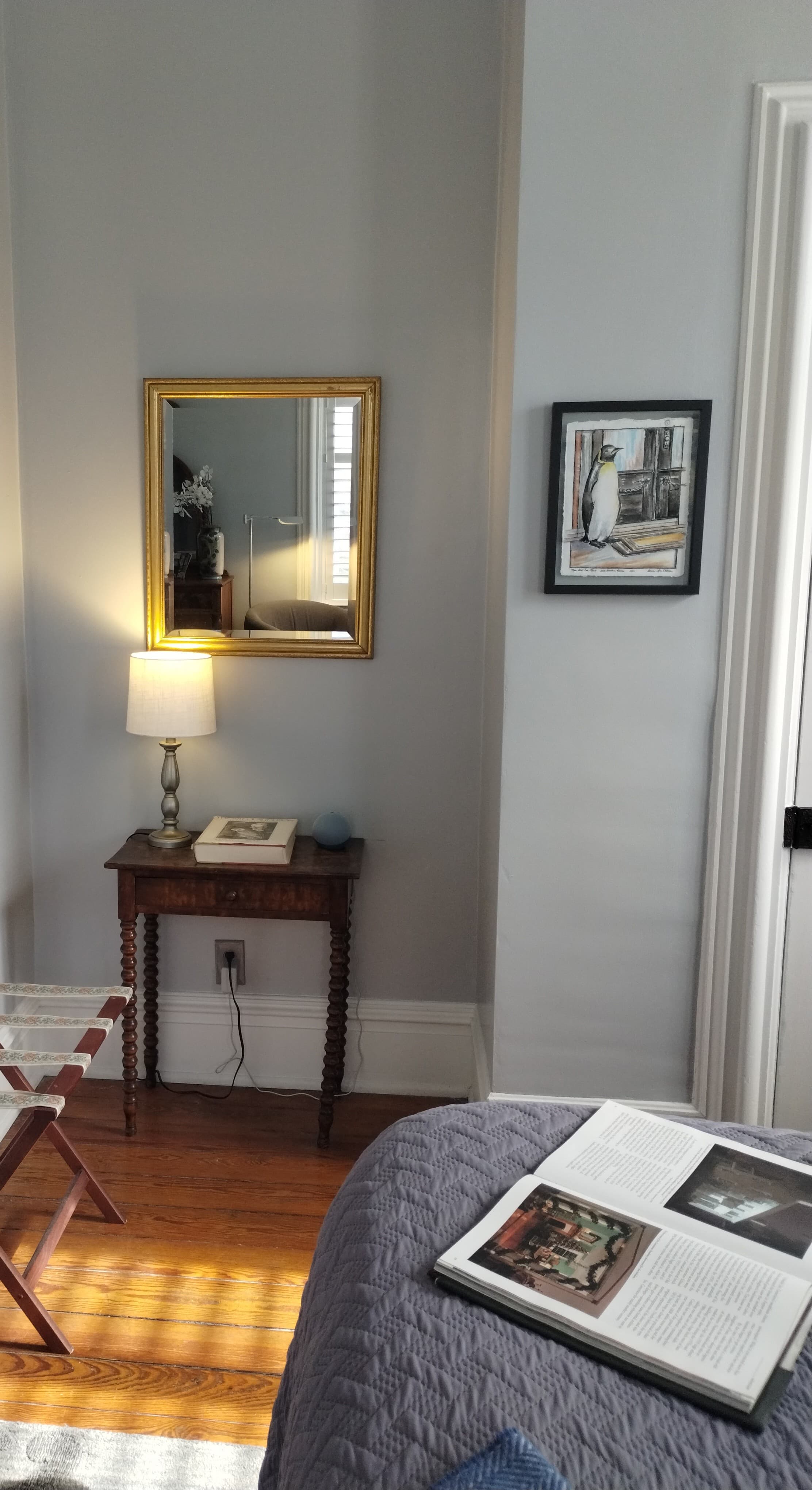 A bedroom with light gray walls featuring a gold-framed rectangular mirror, a small dark wood table with a lamp, a luggage rack, a gray quilted bedspread with an open book, and sunlight streaming in from a window with a white curtain.