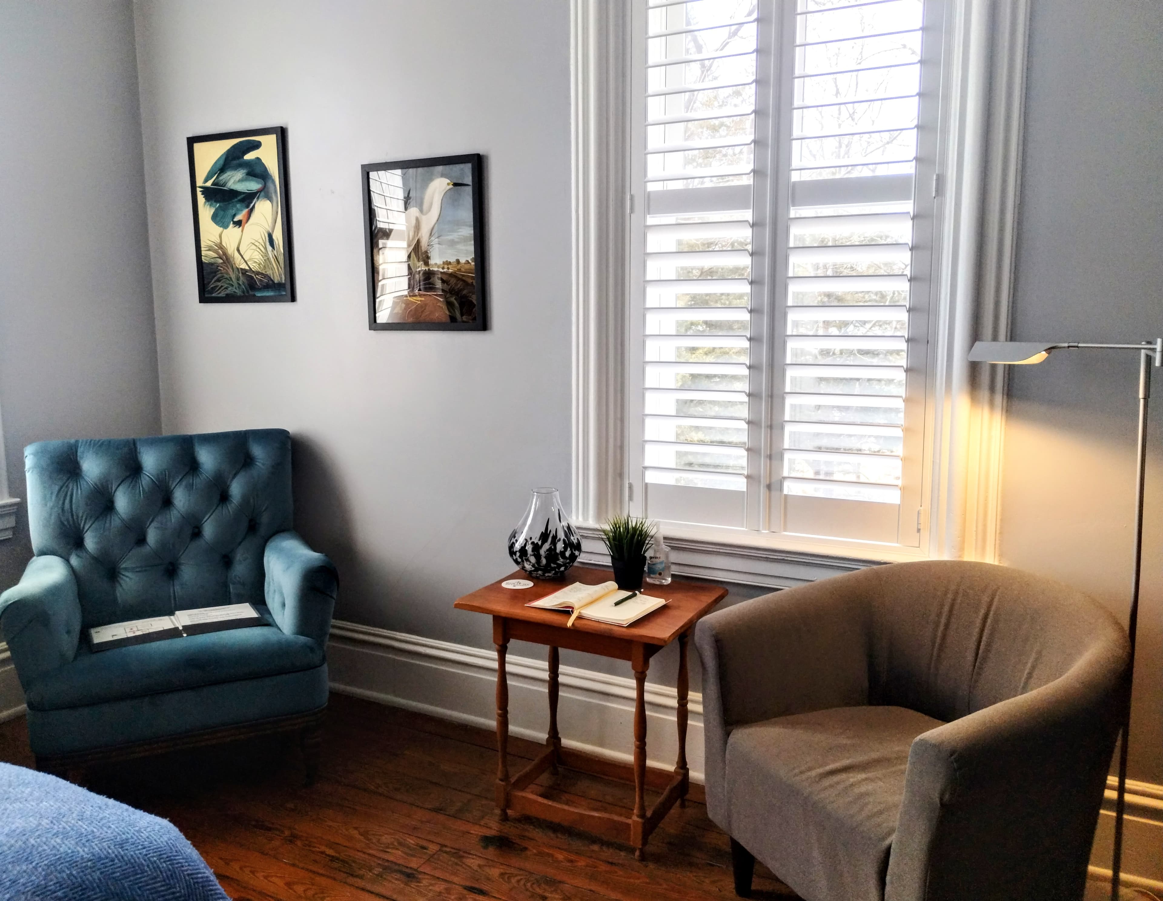 A room with light gray walls featuring a blue tufted armchair, a gray upholstered armchair, a small wooden table with items on top, two framed artworks, and a window with white shutters. Hardwood floors are visible.