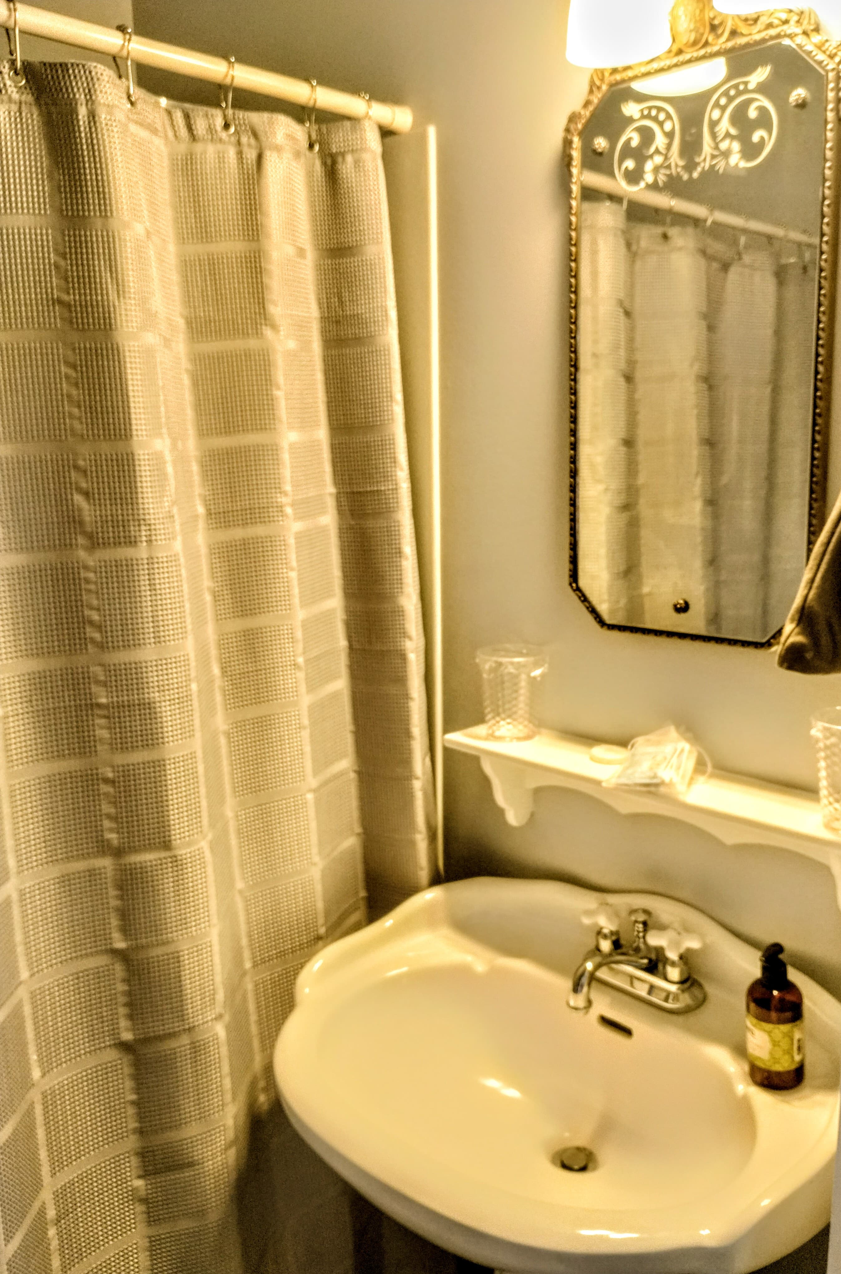 A vintage-style bathroom with a white pedestal sink, a beige shower curtain with a grid pattern, a decorative mirror with an ornate top, a small shelf, and a bottle on the sink.