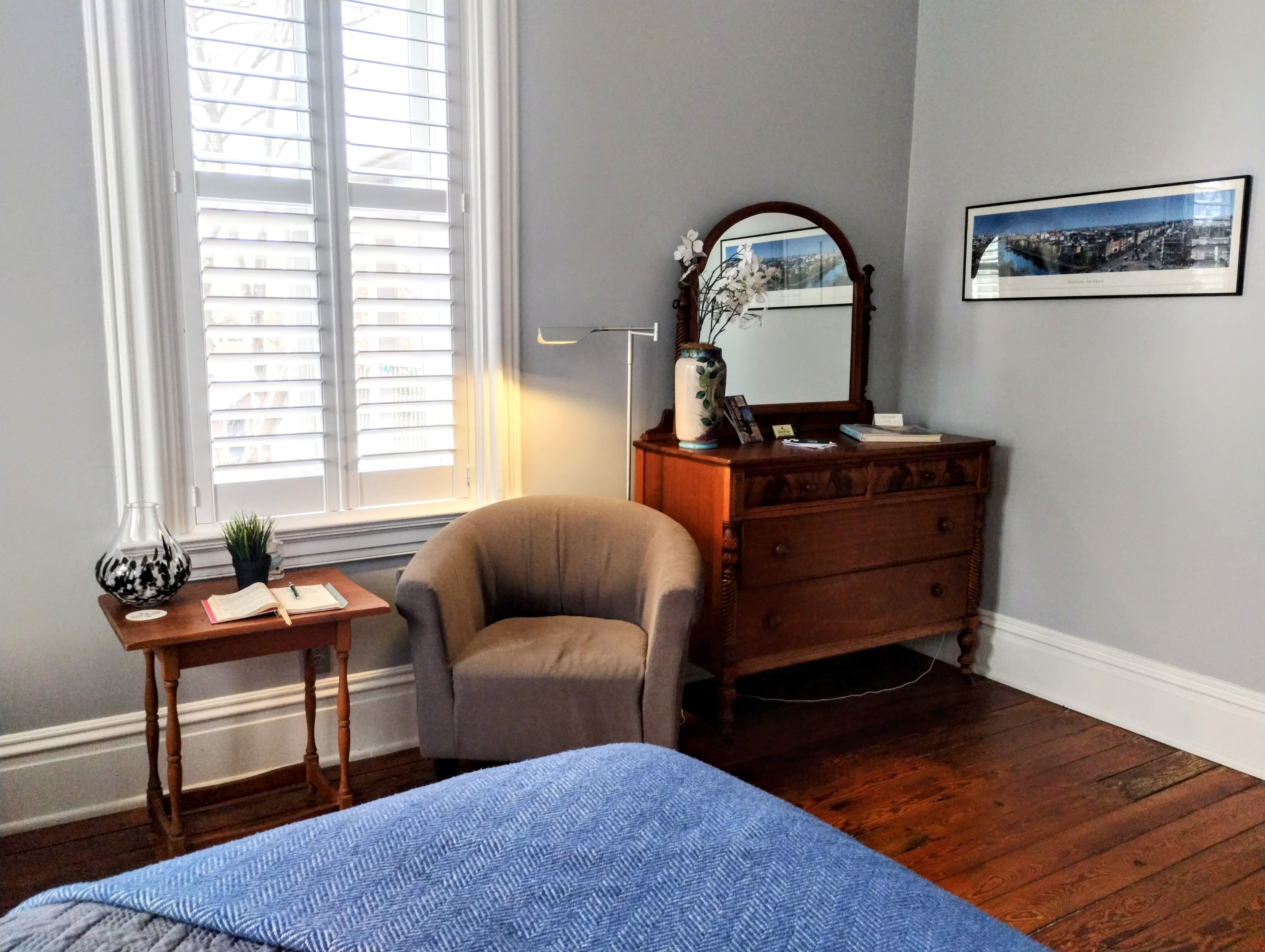A room with a gray armchair, a wooden dresser with a mirror, and a window with white shutters.