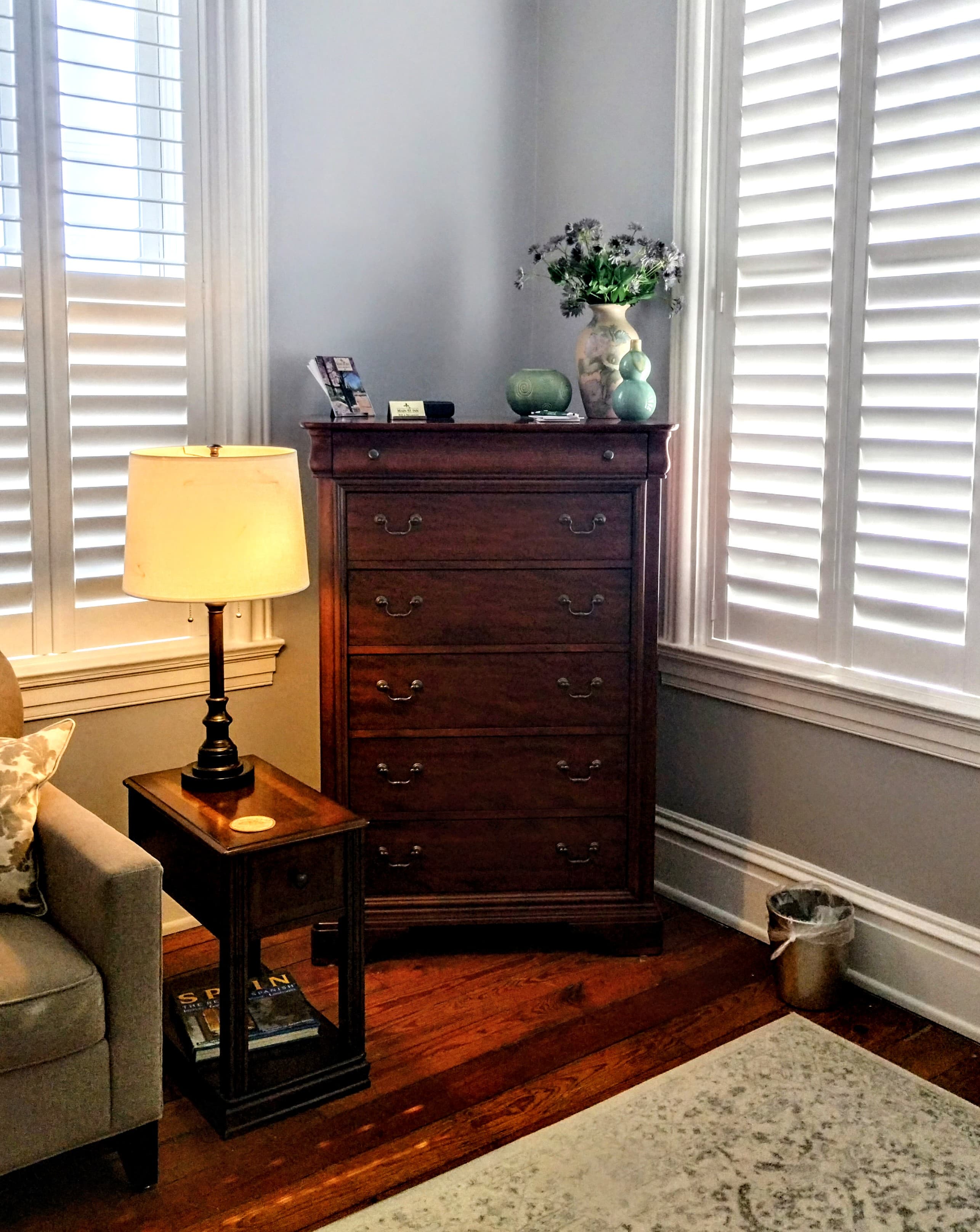 A room with hardwood floors and light gray walls featuring a dark wooden dresser with a vase of flowers, two windows with white slatted shutters, a lamp with a beige shade on a dark wood side table, a beige armchair, and a light-colored rug.
