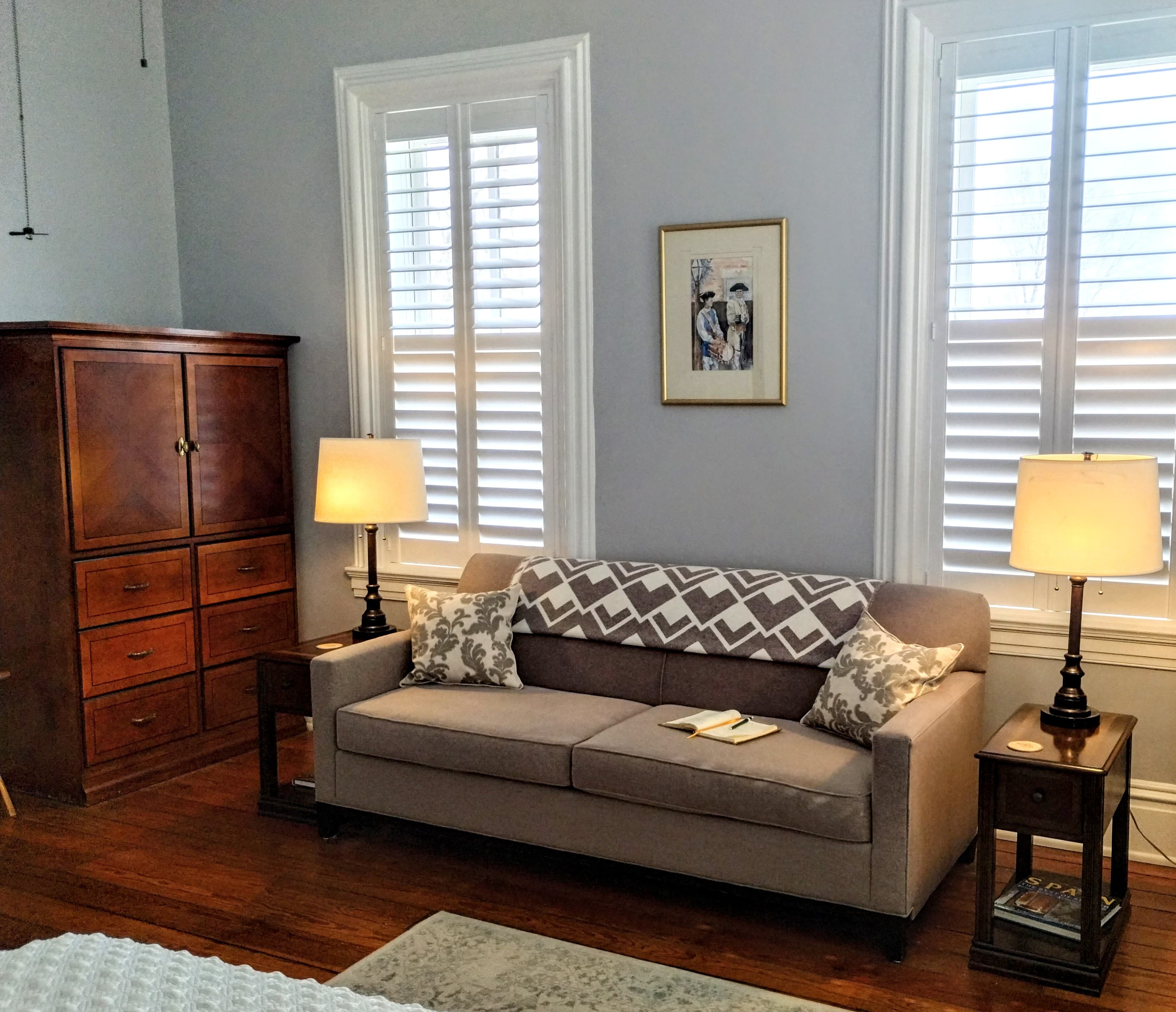 Sitting area with a gray couch, two lamps, a wooden cabinet, framed artwork, and two windows with white shutters.