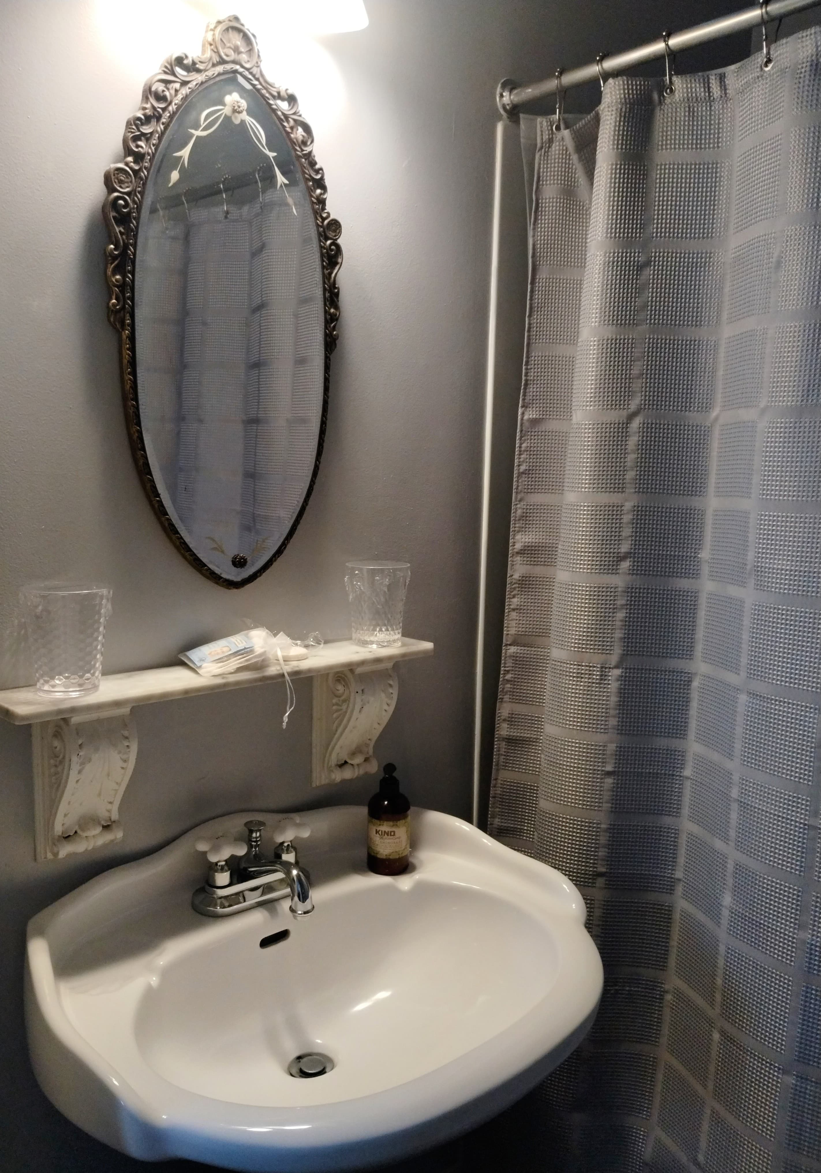 A small bathroom with gray walls featuring a white pedestal sink, an oval mirror with an ornate frame, a small shelf with items, and a shower curtain with a grid pattern.