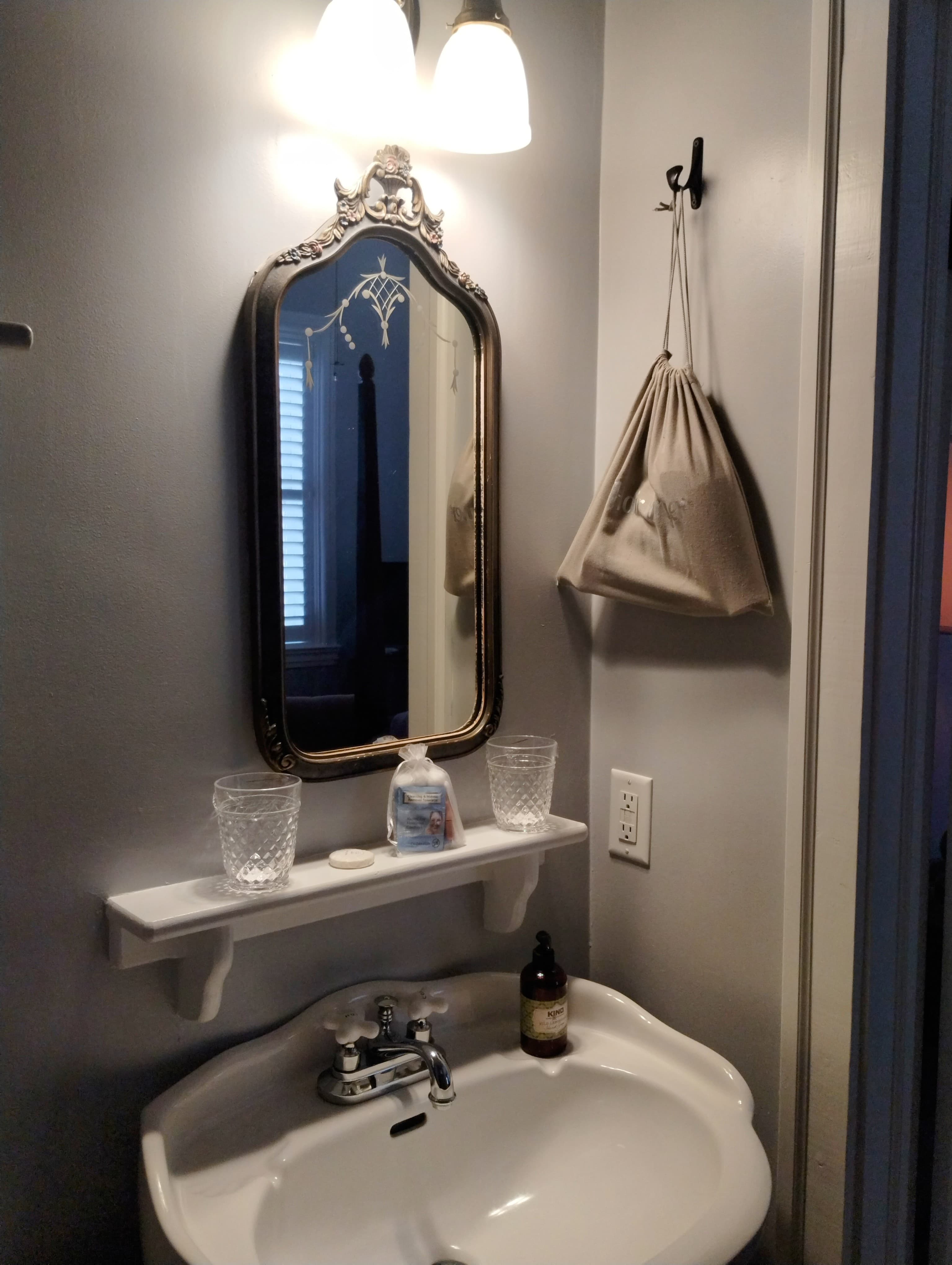 A small bathroom with gray walls featuring a white pedestal sink, a dark ornate mirror, a small white shelf with glasses and a bottle, a hanging bag, and a light fixture with two bulbs.