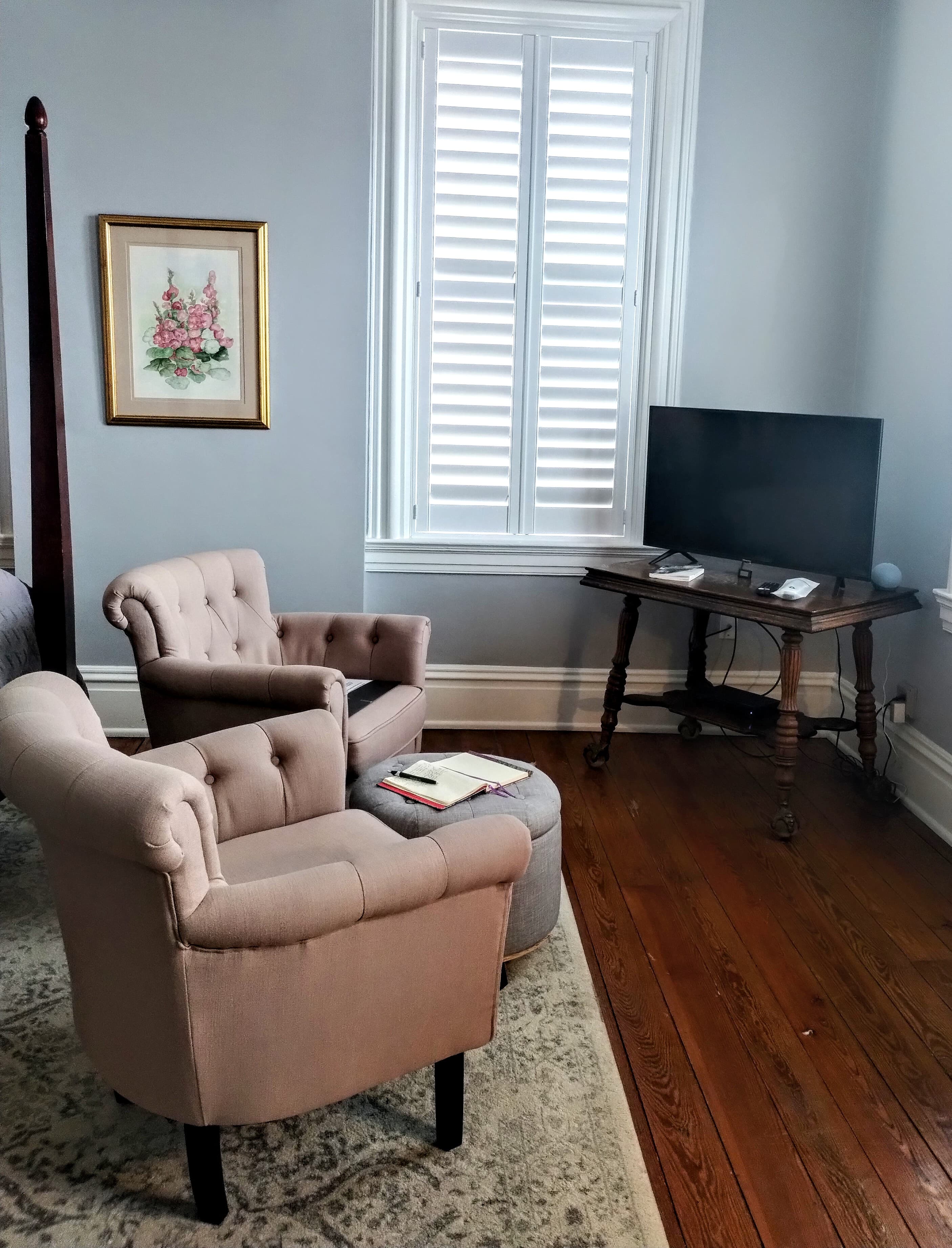 A room with two beige armchairs, a TV on a table, a window with shutters, and framed artwork.