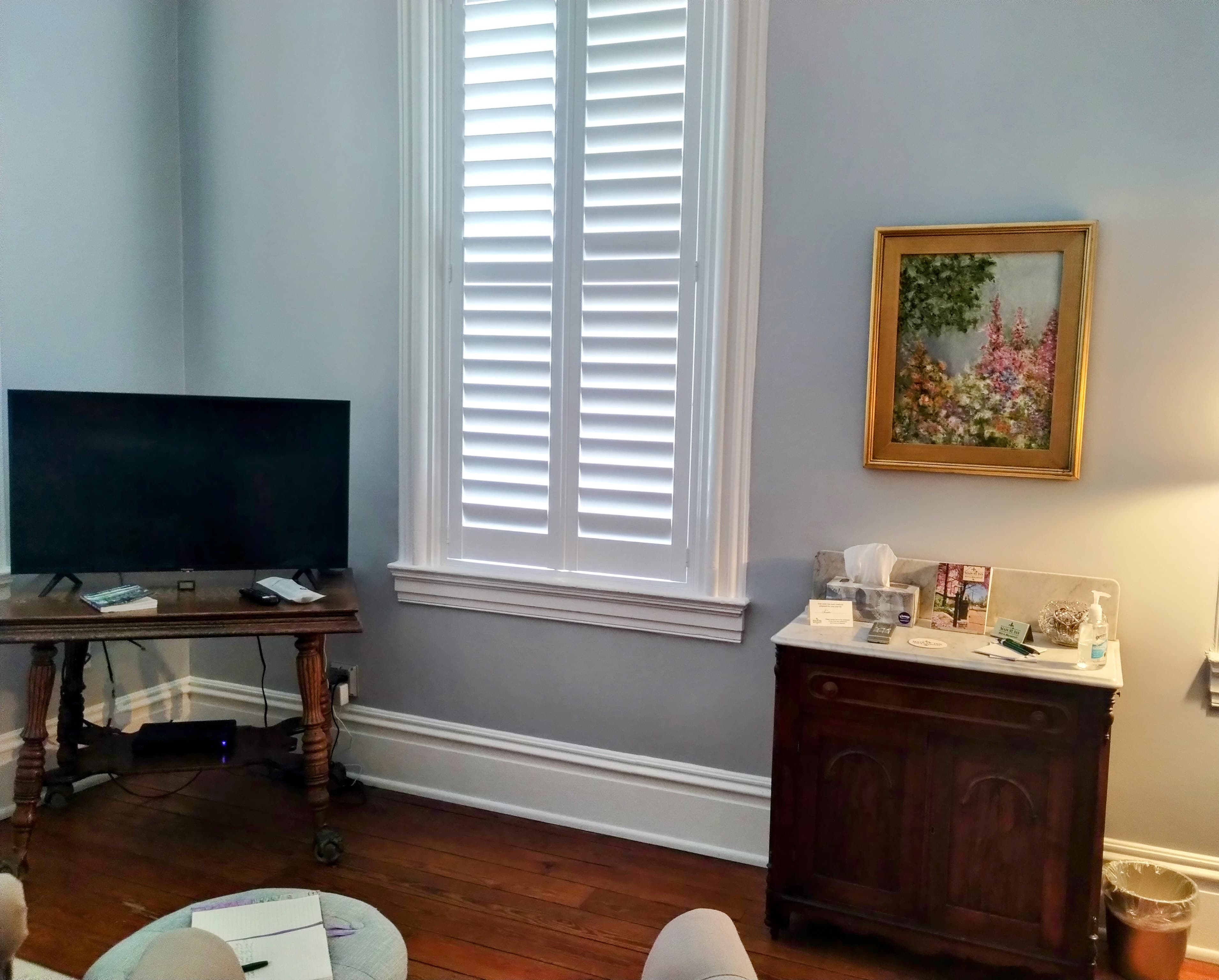 A room with a TV, framed artwork, a dark wood cabinet, and a window with white plantation shutters.