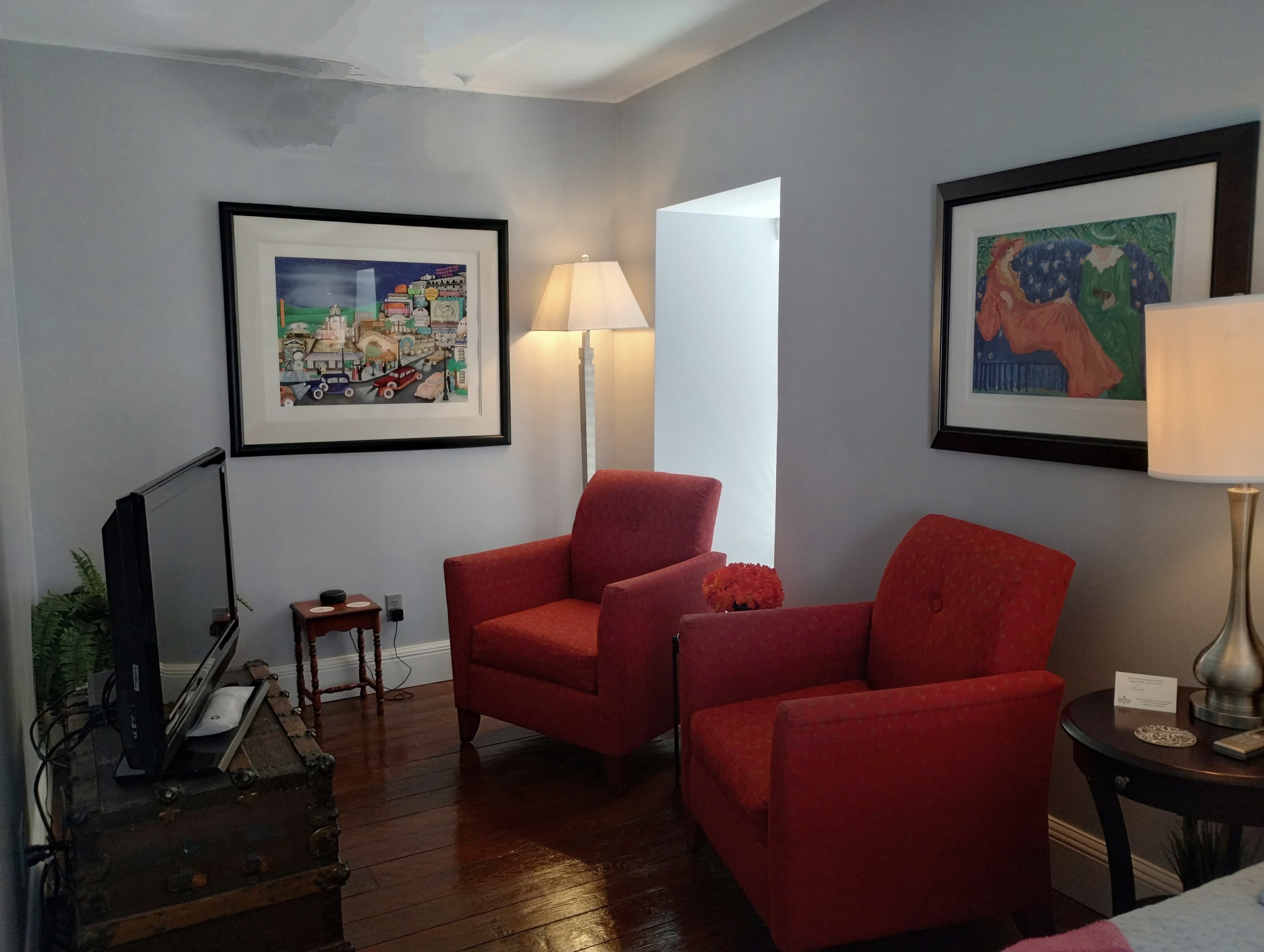 A sitting area with light gray walls featuring two red armchairs, a framed painting with a colorful scene, a floor lamp with a beige shade, a small table between the chairs, and a black television on the left. An air conditioning unit is visible near the ceiling.