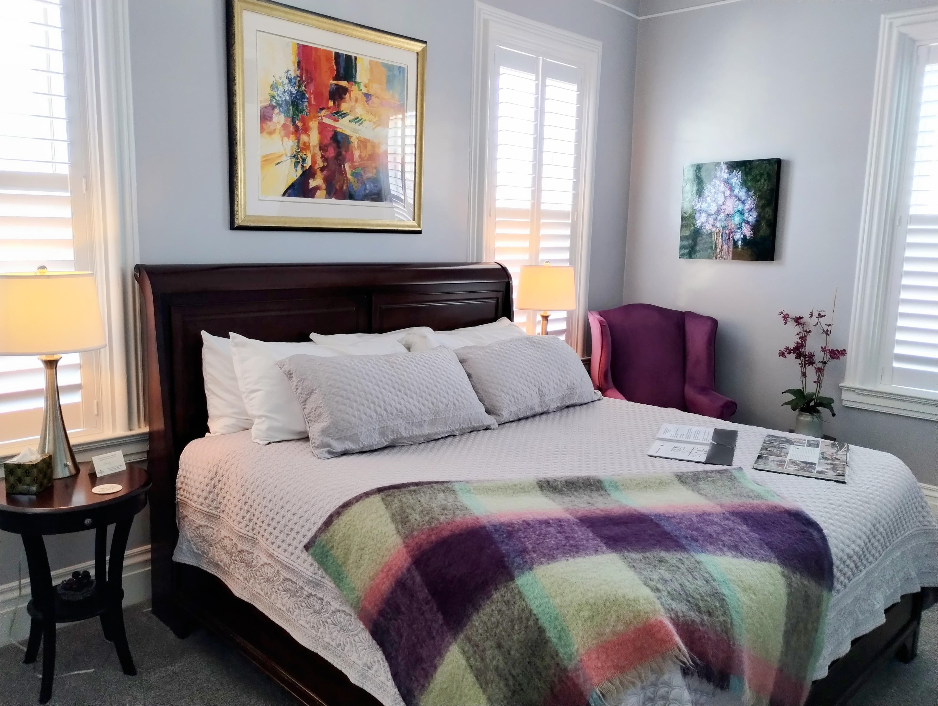 A bright bedroom with light gray walls featuring a dark wood bed with white linens and a colorful plaid throw, two framed artworks on the walls, a small table with a lamp, and a purple armchair in the background.
