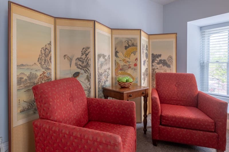 Two red armchairs with a small wooden table between them, a window with a view of trees and a blue sky, and a decorative folding screen with Asian-inspired artwork in the background.