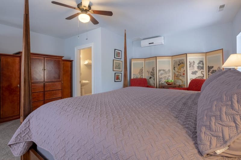 A spacious bedroom with light gray walls featuring a bed with white linens, a dark wood dresser, two bedside tables with lamps, a blue rug with a floral pattern, a ceiling fan, and a doorway leading to a bathroom.
