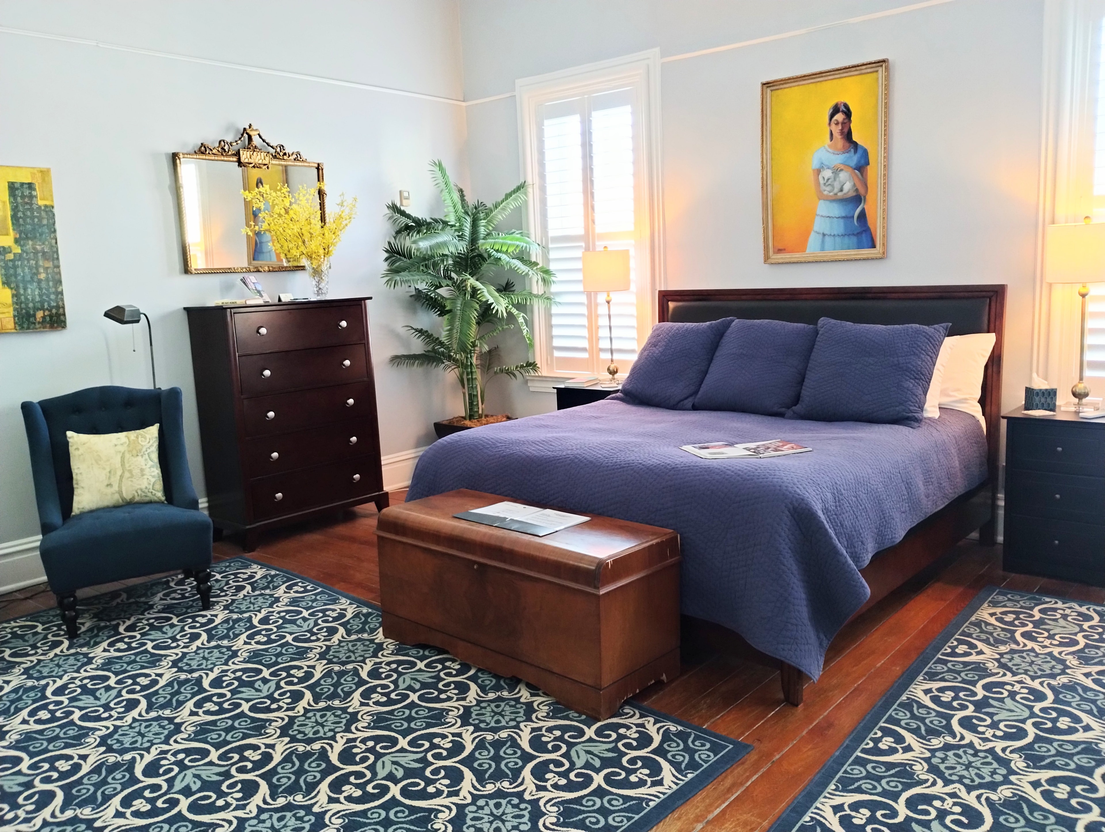A bedroom with a blue bedspread, a wooden dresser, a blue armchair, and a patterned rug.