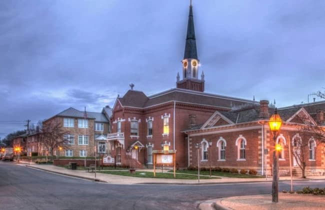 A brick church with a tall steeple is illuminated at dusk alongside a quiet street.