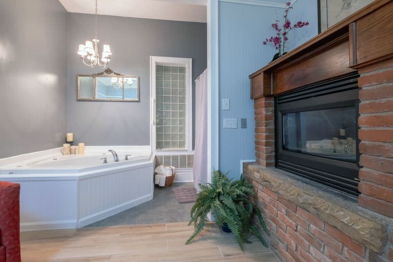 A room featuring a white corner jetted bathtub, a brick fireplace with a wooden mantel, light wood flooring, a decorative chandelier, and a glimpse of a red armchair.