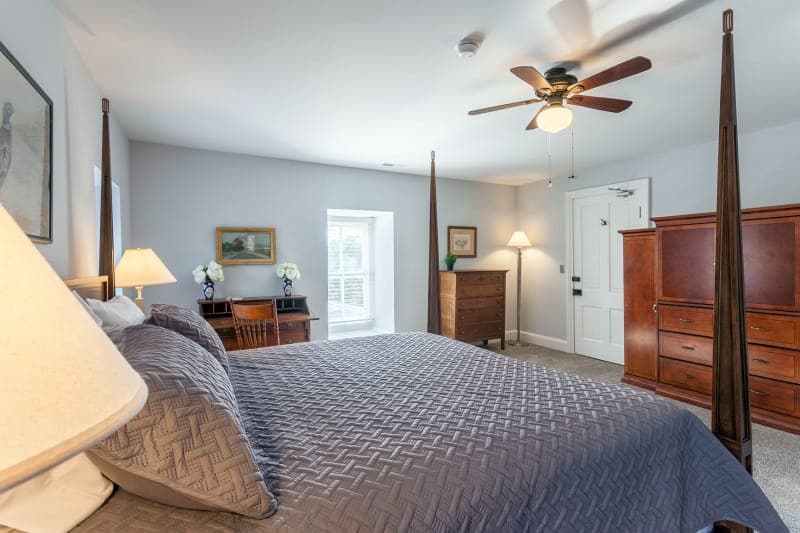 A spacious bedroom with light gray walls featuring a bed with white linens, a dark wood dresser, two bedside tables with lamps, a blue rug with a floral pattern, a ceiling fan, and a doorway leading to a bathroom.