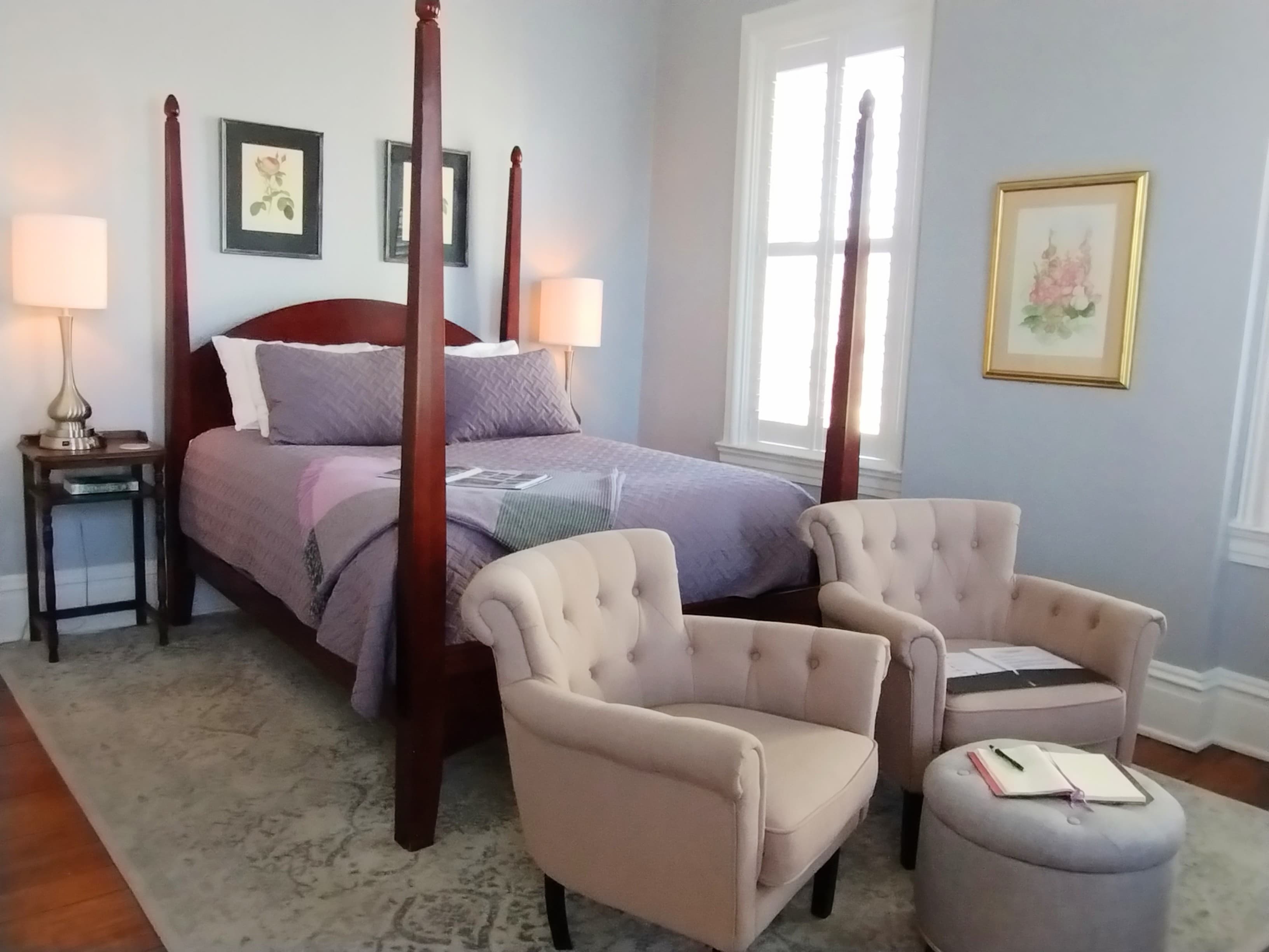 A bedroom with light blue walls featuring a dark wood four-poster bed with purple bedding, two beige tufted armchairs with a matching ottoman, two framed artworks, two bedside lamps on a small side table, and a light-colored rug.