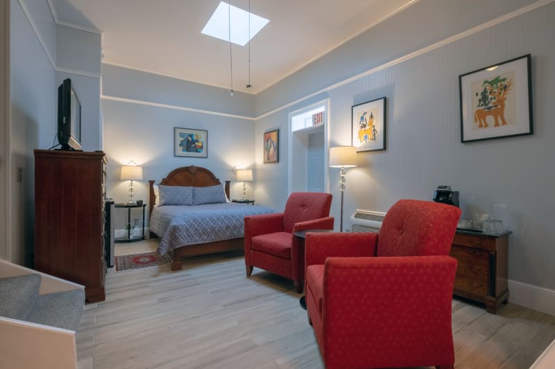 A well-lit bedroom with light gray walls and two bright red armchairs facing a wooden bed with blue bedding. Artwork hangs on the walls, and a skylight provides natural light.