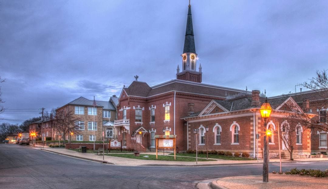 A brick church with a tall steeple is illuminated by street lamps on a quiet street at dusk.