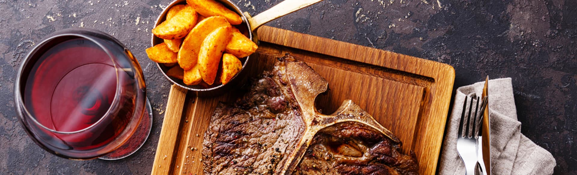 A wooden platter with a large steak, a side of potato wedges in a cup, and a glass of red wine.
