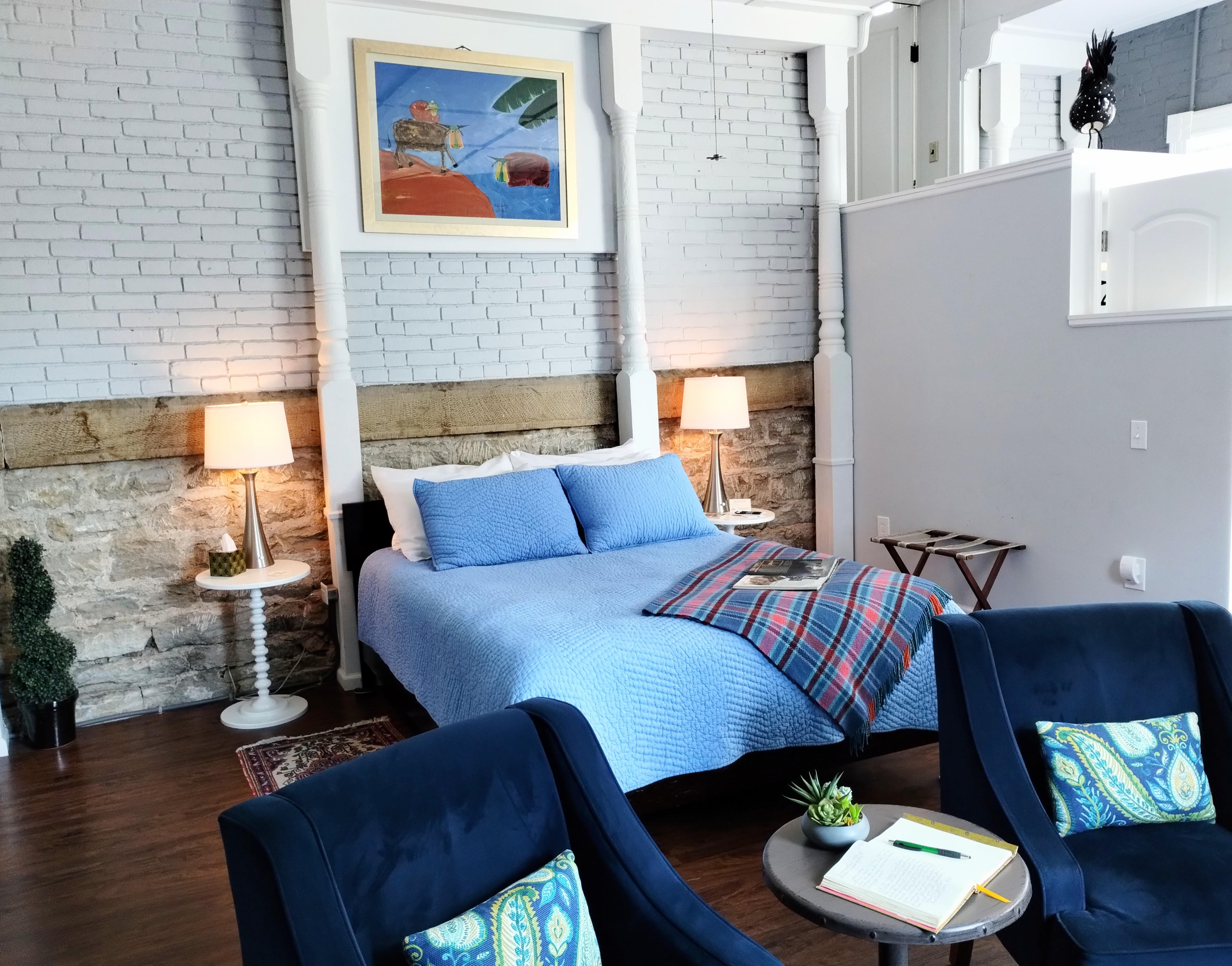 A bedroom with exposed brick walls, a blue bed with a patterned throw, two dark blue armchairs with patterned pillows, two bedside lamps, a small table between the chairs, and a framed painting above the bed.