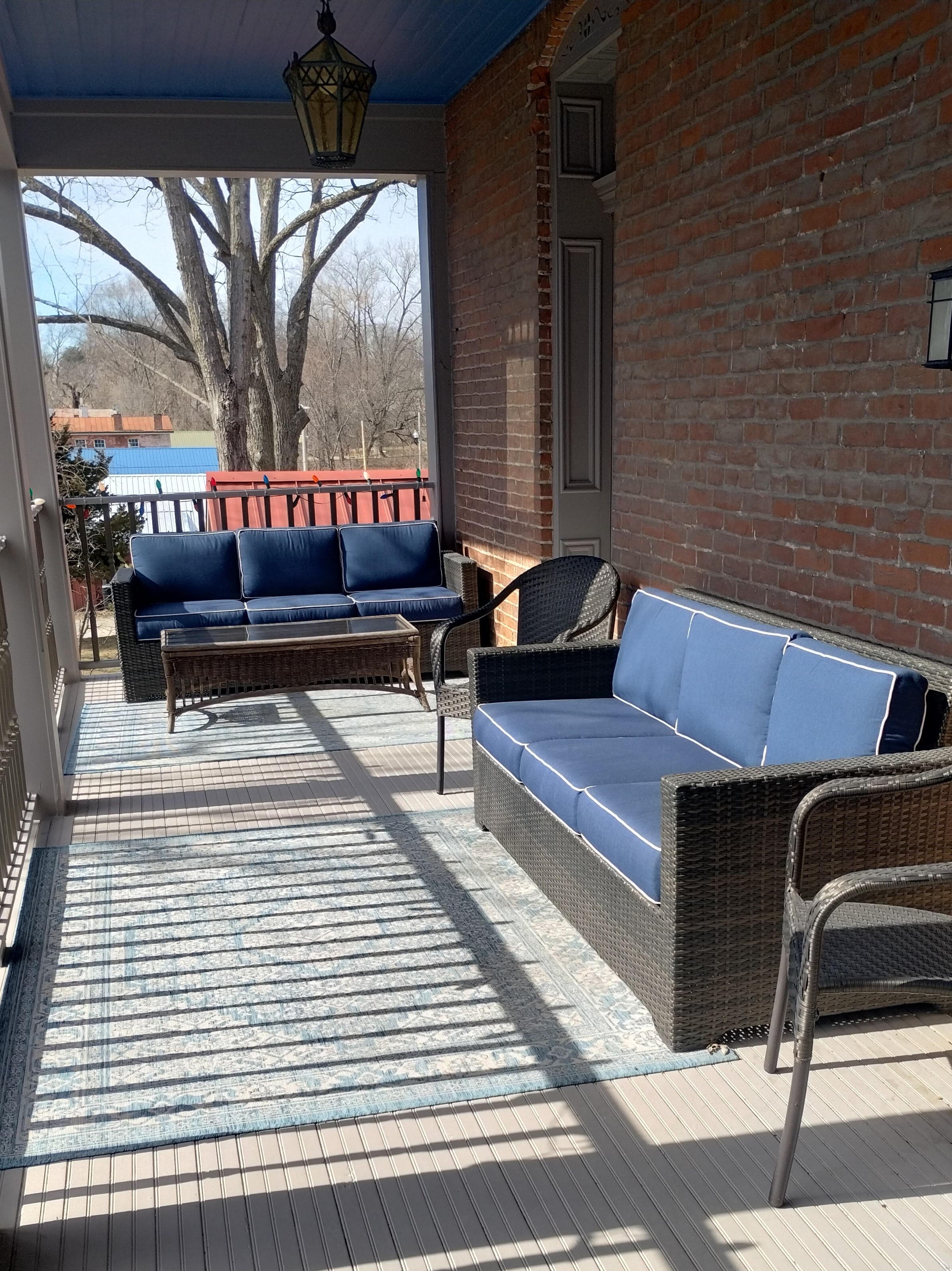 An outdoor porch with a blue ceiling, a brick wall on one side, and two dark-colored couches with blue cushions. A glass-topped table sits between the couches. A large tree and a glimpse of a colorful building are visible in the background.