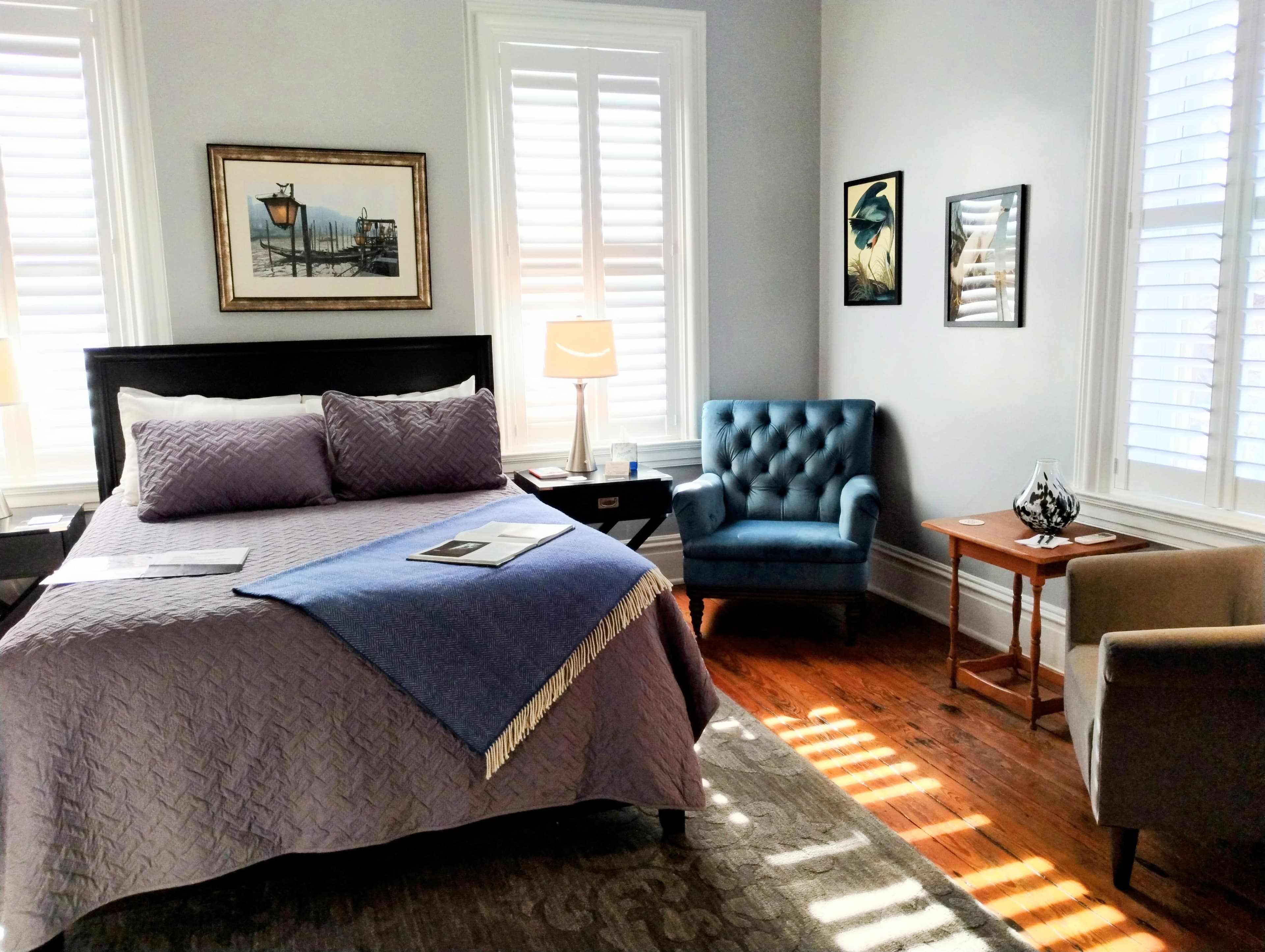 A bright bedroom with light gray walls featuring a dark wood bed with gray bedding and a blue throw, a tufted blue armchair, two framed artworks, two small side tables, and sunlight streaming through white shuttered windows onto hardwood floors.