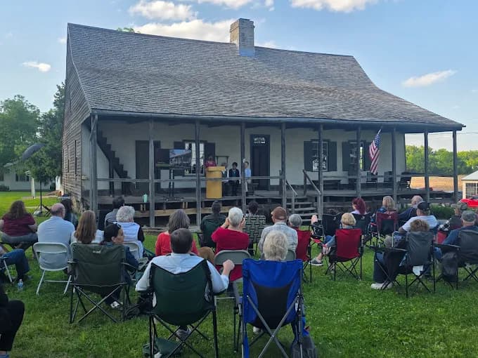 A crowd sits in front of a historic house while observing a presentation on its porch.
