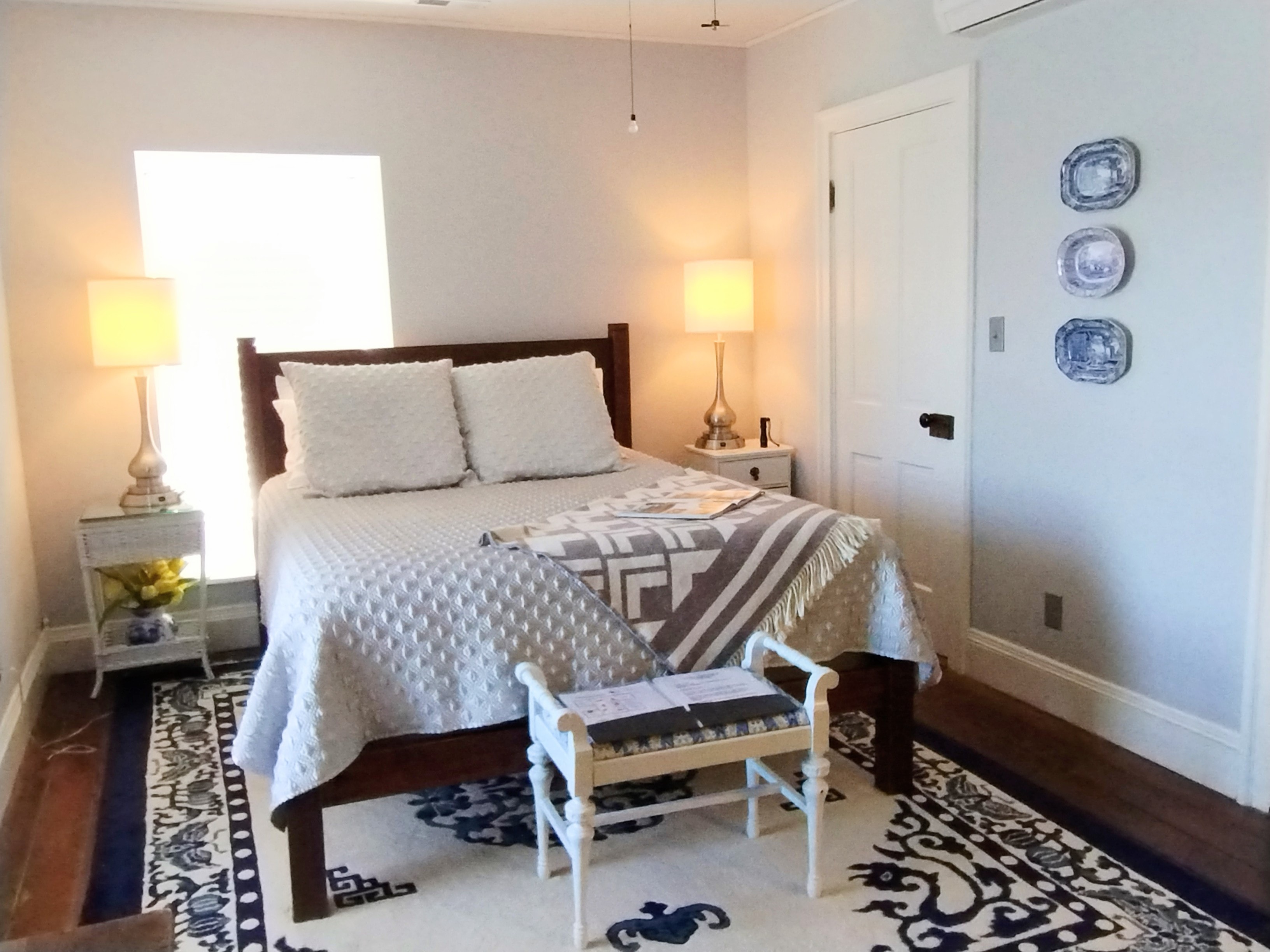 A spacious bedroom with light gray walls featuring a bed with white linens, a dark wood dresser with a mirror, two bedside tables with lamps, a blue rug with a floral pattern, a ceiling fan, and a doorway leading to a bathroom.