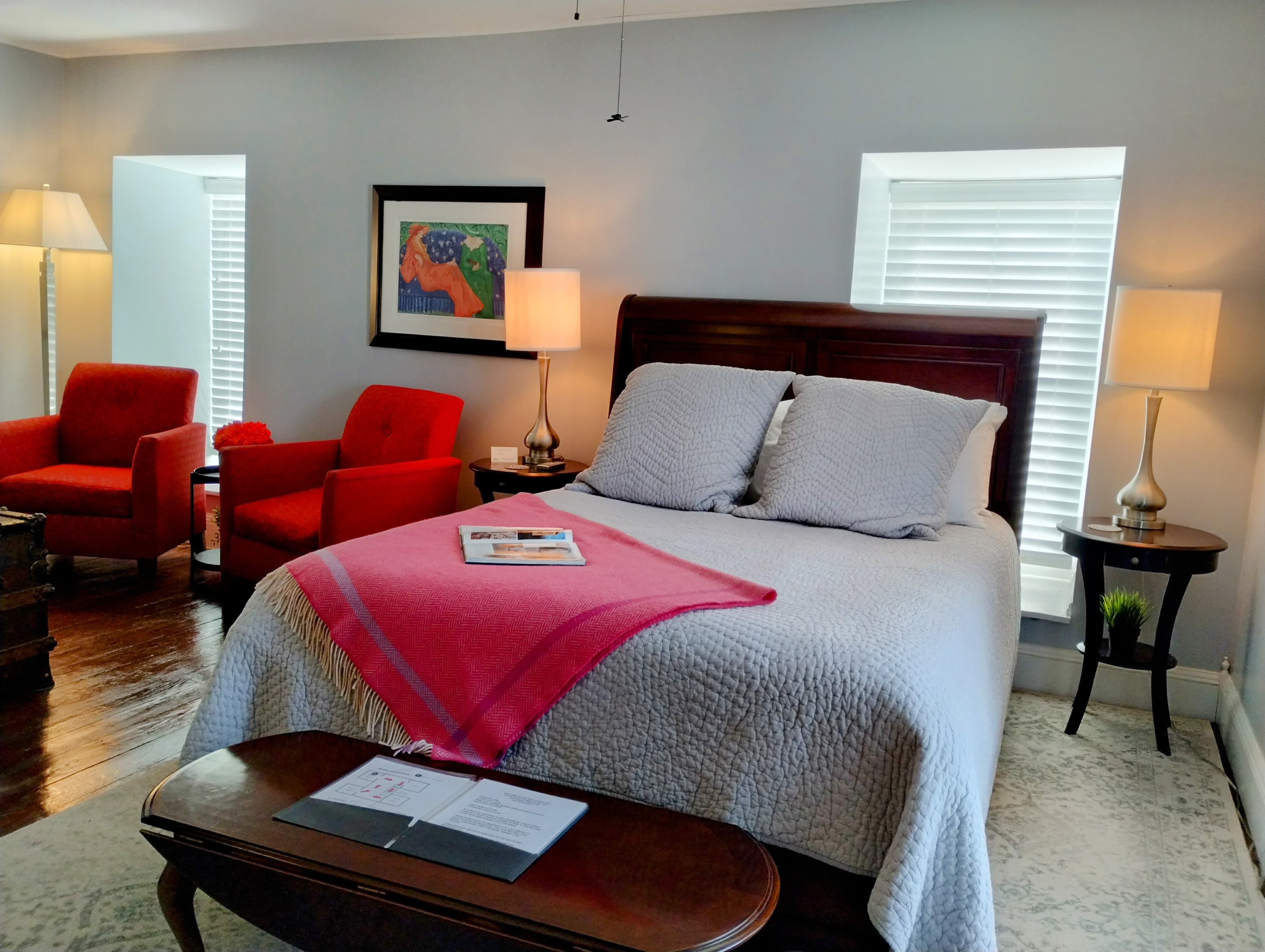 A bedroom with light gray walls featuring a dark wood bed with white linens and a gray throw, a red armchair, two bedside lamps on small tables, a dark wood bench at the foot of the bed, a ceiling fan, and a window with a deep sill.