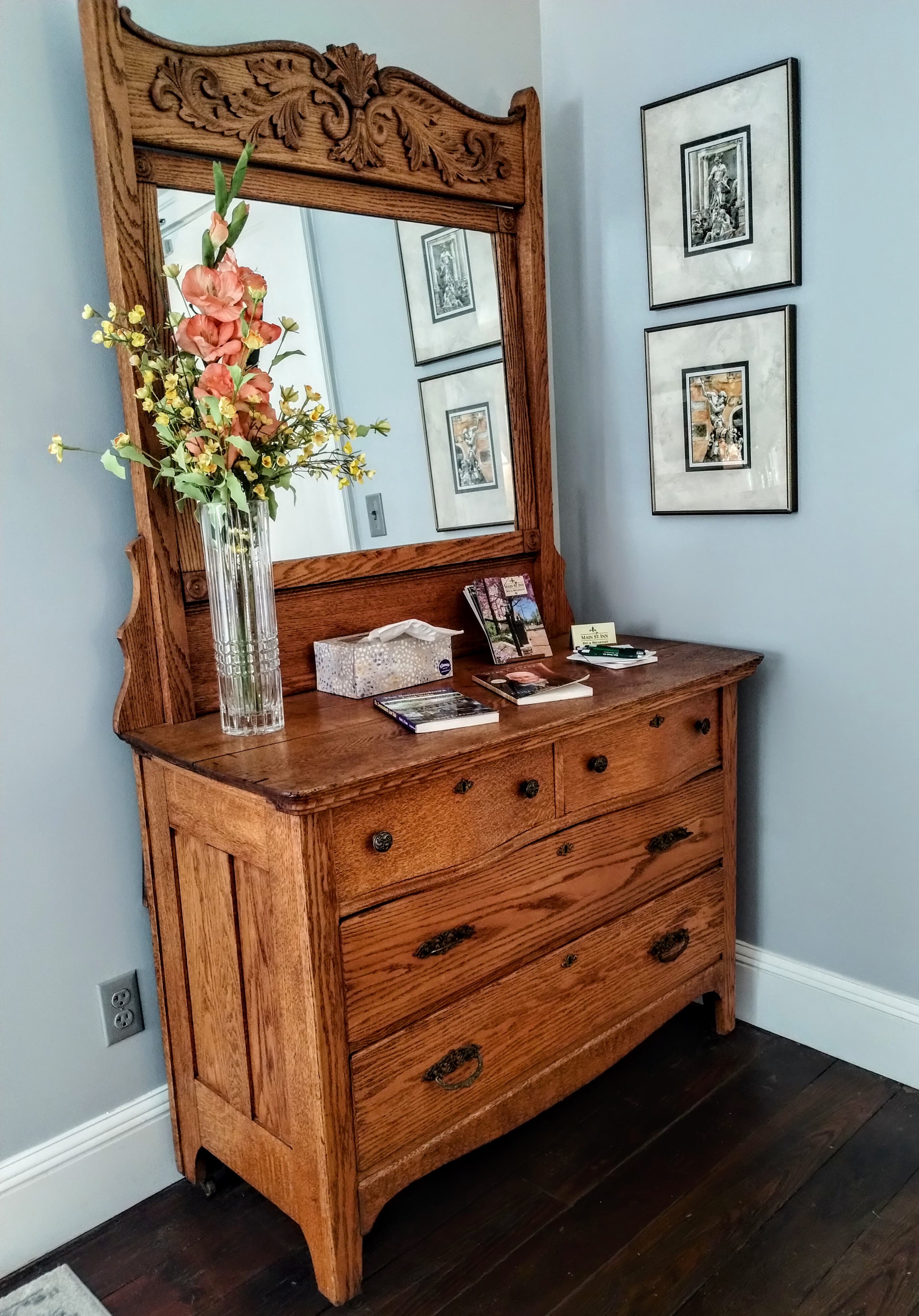 A vintage wooden dresser with a large ornate mirror, adorned with a vase of flowers and various items on top, set against a light blue wall.