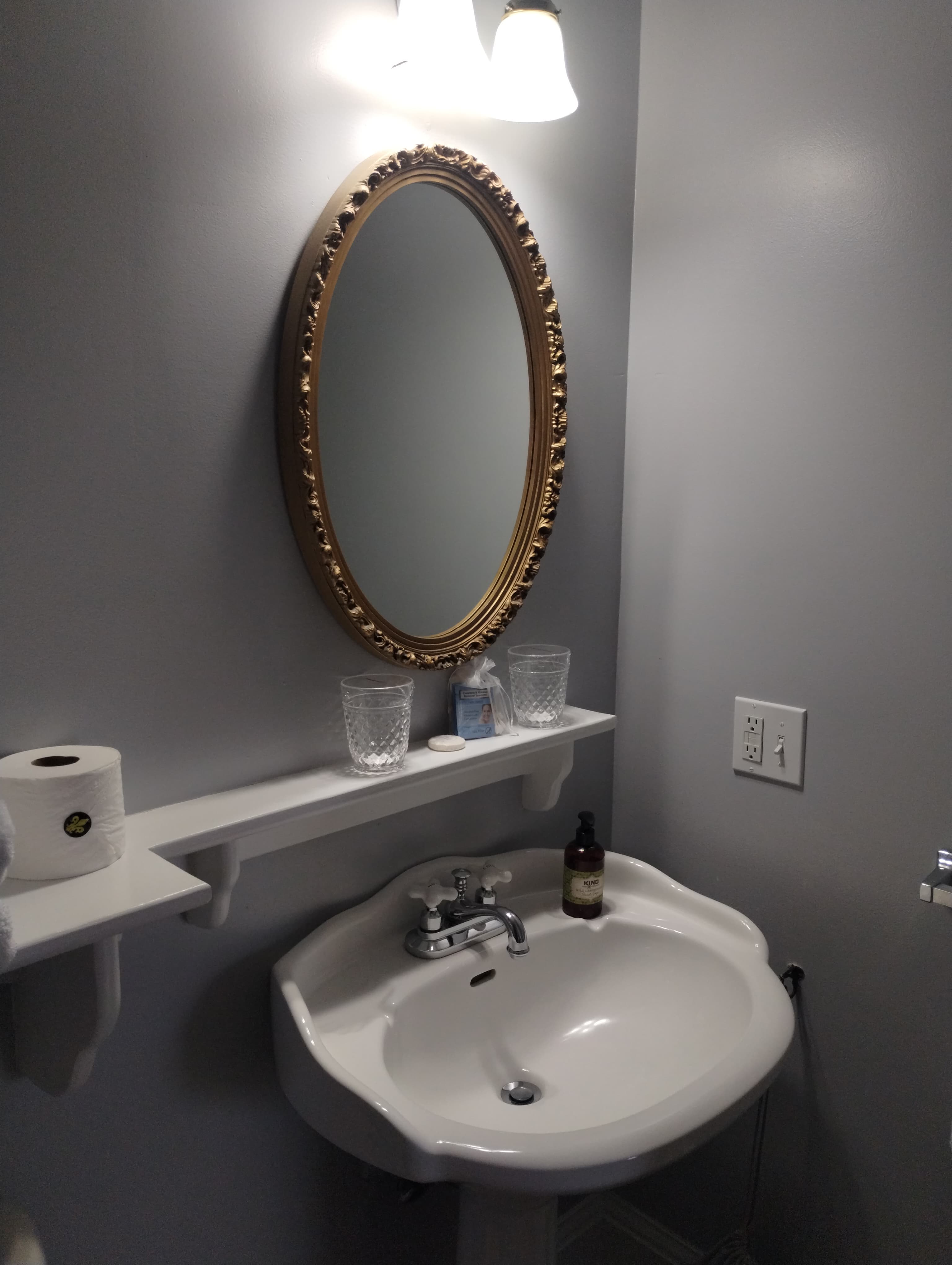 A bathroom with a round mirror, a sink, and a shelf holding toiletries and a toilet paper roll.