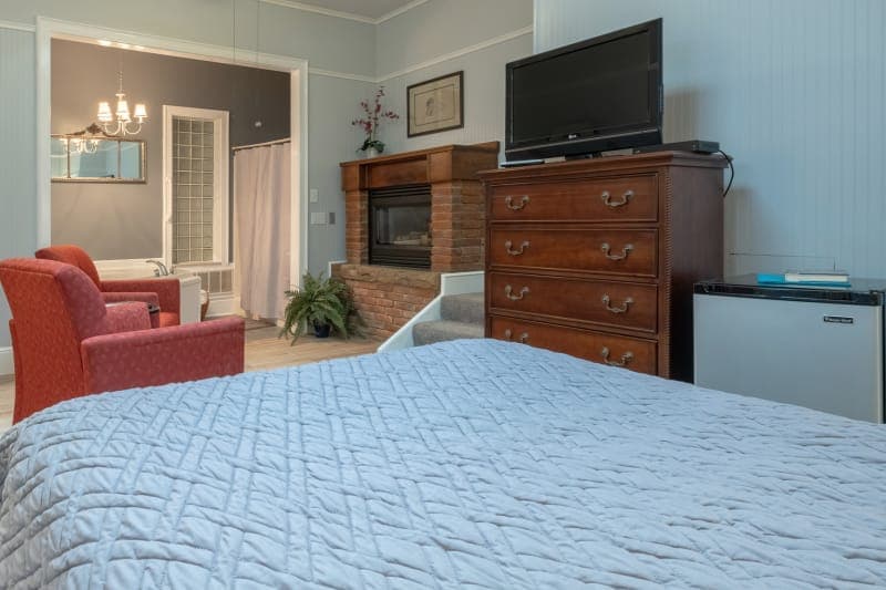 A bedroom with a striped comforter on the bed, a wooden dresser with a TV, a red armchair, and a glimpse into an adjacent room.