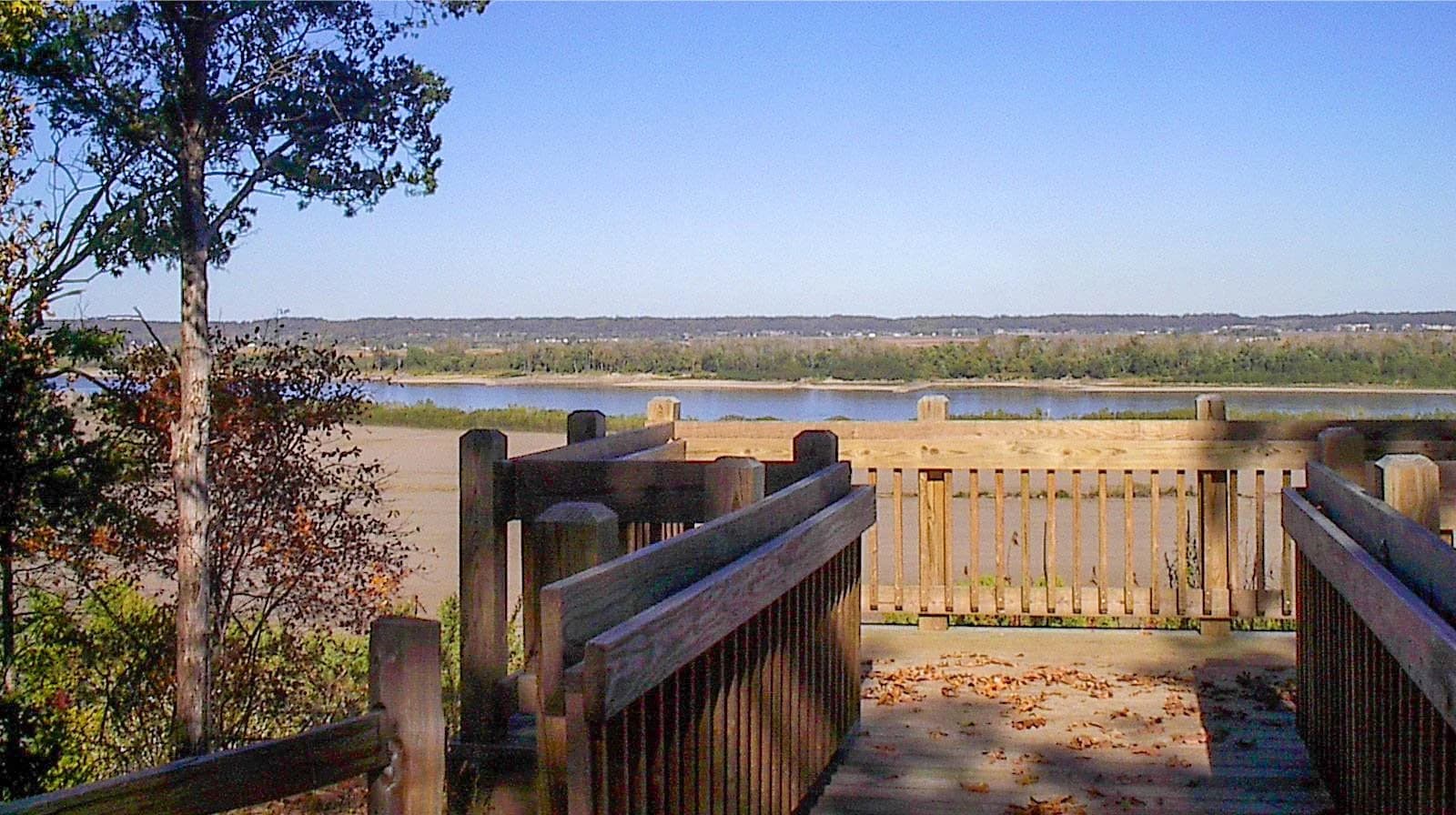 View from a wooden overlook of a river surrounded by trees and rolling hills under a clear blue sky.