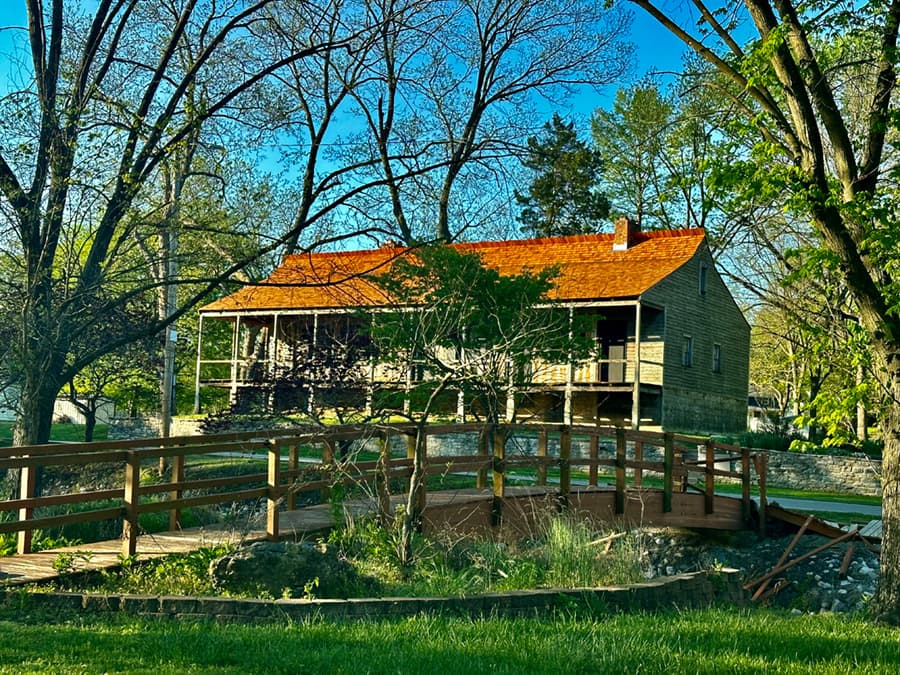 Historic wooden house with a red roof surrounded by trees and a wooden bridge.